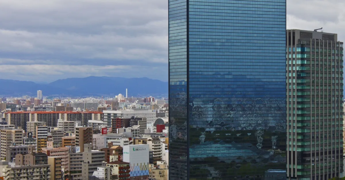 Skyline von Osaka, mit einem riesigen Hochhaus und im Hintergrund niedrigere Häuser und eine Bergkette