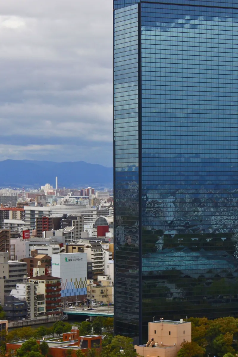 Skyline von Osaka, mit einem riesigen Hochhaus und im Hintergrund niedrigere Häuser und eine Bergkette