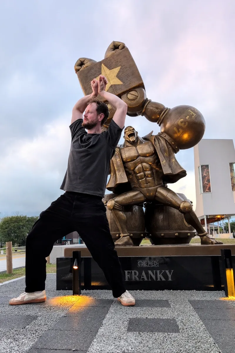 Muskulöser Mann posiert vor der Franky-Statue am Bahnhof Takamori