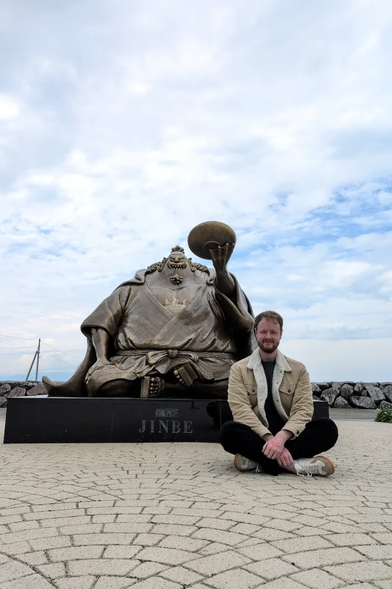 Mann sitzt im Schneidersitz neben der Statue von Jinbe an der Nagabeta Seabed Road, Uto