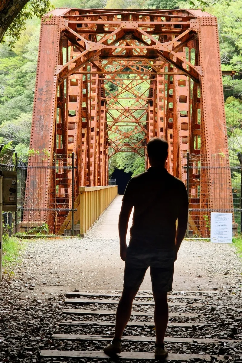 Mann steht im Schatten im Tunnelausgang eines einem verlassenen Eisenbahntunnels, der auf eine rostrote Stahlbrücke zuführt in einem Wald auf dem Takedao Abandoned Railway Hike