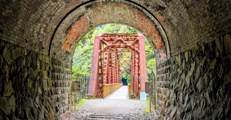 Blick aus einem verlassenen Eisenbahntunnel auf eine rostrote Stahlbrücke in einem Wald auf dem Takedao Abandoned Railway Hike