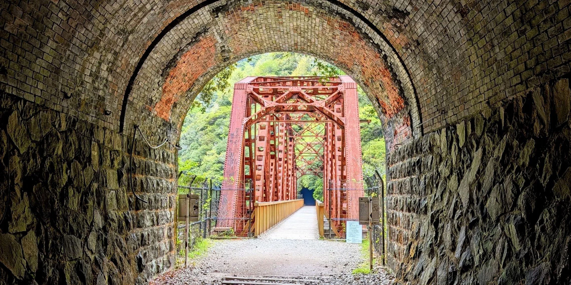 Blick aus einem verlassenen Eisenbahntunnel auf eine rostrote Stahlbrücke in einem Wald auf dem Takedao Abandoned Railway Hike